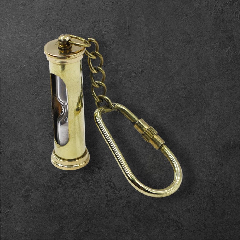 Puede incluir: Un llavero de reloj de arena de lat&oacute;n con una peque&ntilde;a cadena y un mosquet&oacute;n grande. El reloj de arena tiene forma cil&iacute;ndrica con una secci&oacute;n de vidrio transparente para ver la arena. El metal tiene un acabado pulido y brillante.