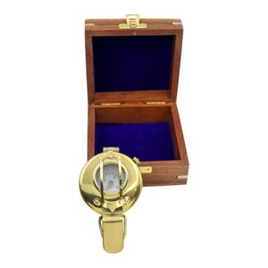 May include: A polished brass pocket compass with a hinged lid and a circular face featuring cardinal directions, positioned in front of an open square wooden storage box with a deep purple velvet interior lining and a gold-toned metal latch.