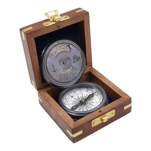 May include: A vintage compass and calendar set in a wooden box. The box is open, revealing a compass with a white face and a calendar dial. The box is made of dark wood with brass accents and a blue velvet interior.