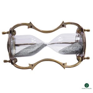 Puede incluir: Un reloj de arena con marco de lat&oacute;n met&aacute;lico, soportes curvos y cuatro patas peque&ntilde;as. El bulbo de vidrio transparente contiene arena gris fina. El marco tiene un dise&ntilde;o decorativo con acentos puntiagudos. El logo Dollond London es visible.