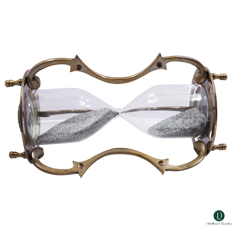Puede incluir: Un reloj de arena con marco de lat&oacute;n met&aacute;lico, soportes curvos y cuatro patas peque&ntilde;as. El bulbo de vidrio transparente contiene arena gris fina. El marco tiene un dise&ntilde;o decorativo con acentos puntiagudos. El logo Dollond London es visible.