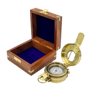 May include: A polished brass nautical compass with a hinged lid and a white dial featuring cardinal directions, positioned next to an open square wooden storage box with brass corner accents and a deep blue velvet interior lining.