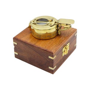 May include: A polished brass nautical compass with a hinged lid and directional markings sits atop a square, dark brown wooden box. The box features brass corner accents and a central brass latch mechanism on the front side for secure closure.