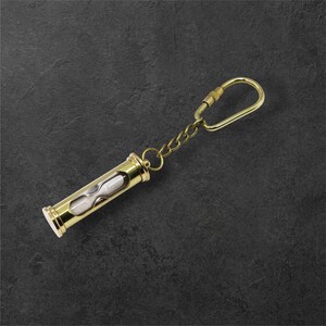 Puede incluir: Un llavero de reloj de arena dorado con una peque&ntilde;a cadena y cierre. El reloj de arena tiene forma cil&iacute;ndrica con arena blanca en su interior. El llavero es de metal y tiene un acabado pulido.