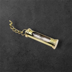 Puede incluir: Un peque&ntilde;o reloj de arena dorado con una cadena. El reloj de arena est&aacute; encerrado en un marco met&aacute;lico cil&iacute;ndrico con un tubo de vidrio transparente en el centro. La arena del interior es blanca. La cadena est&aacute; unida a un anillo en la parte superior.