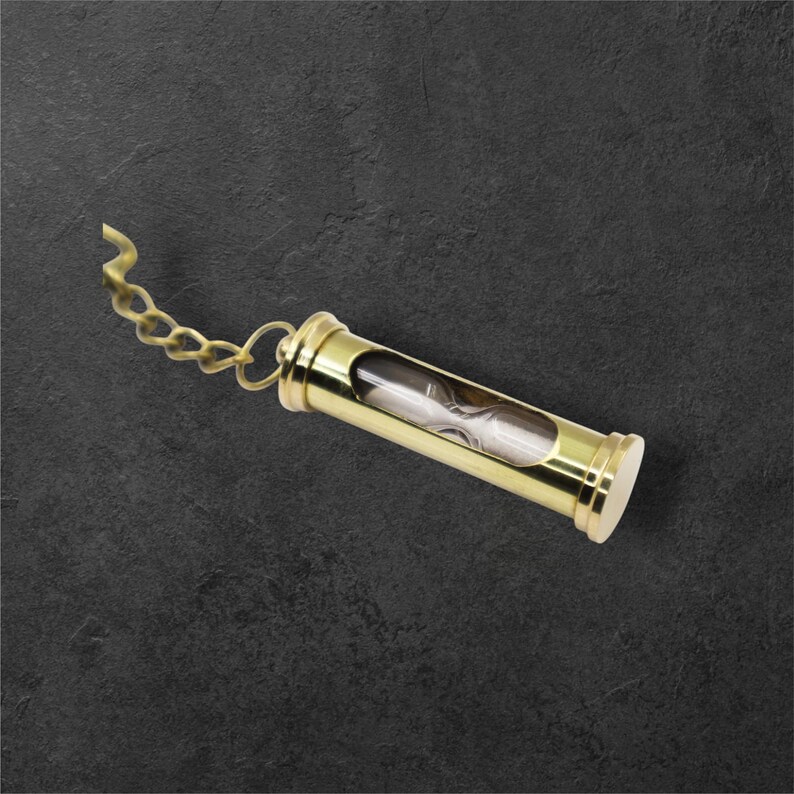 Puede incluir: Un peque&ntilde;o reloj de arena dorado con una cadena. El reloj de arena est&aacute; encerrado en un marco met&aacute;lico cil&iacute;ndrico con un tubo de vidrio transparente en el centro. La arena del interior es blanca. La cadena est&aacute; unida a un anillo en la parte superior.