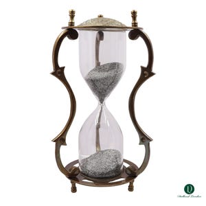 Puede incluir: Un reloj de arena con un bulbo de vidrio transparente lleno de arena gris, encerrado en un marco de metal color bronce oscuro con soportes curvos y una tapa texturizada. La base es circular con pies peque&ntilde;os, el logo Dollond London es visible.
