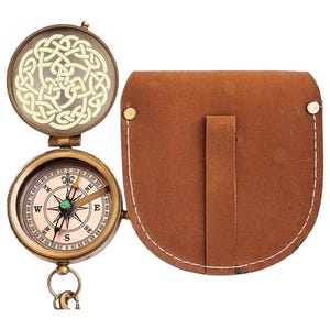May include: A brass pocket compass with a hinged lid featuring an intricate Celtic knot design. The compass face displays cardinal directions, degree markings, and a central needle. A brown leather pouch with white stitching accompanies the item.