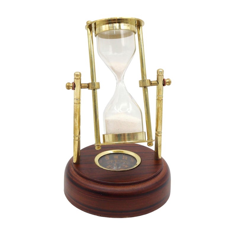 K&ouml;nnte beinhalten: Eine Sanduhr mit einer Glaskugel gef&uuml;llt mit wei&szlig;em Sand, in einem Messingrahmen. Der Rahmen wird von einem dunkelbraunen Holzsockel getragen. Die Sanduhr ist ein dekorativer Artikel.