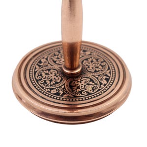 May include: A copper-toned metal stand featuring a circular base with a black inlay. The base is decorated with intricate floral and leaf patterns in copper. A smooth, cylindrical copper-toned post extends vertically from the center of the base.
