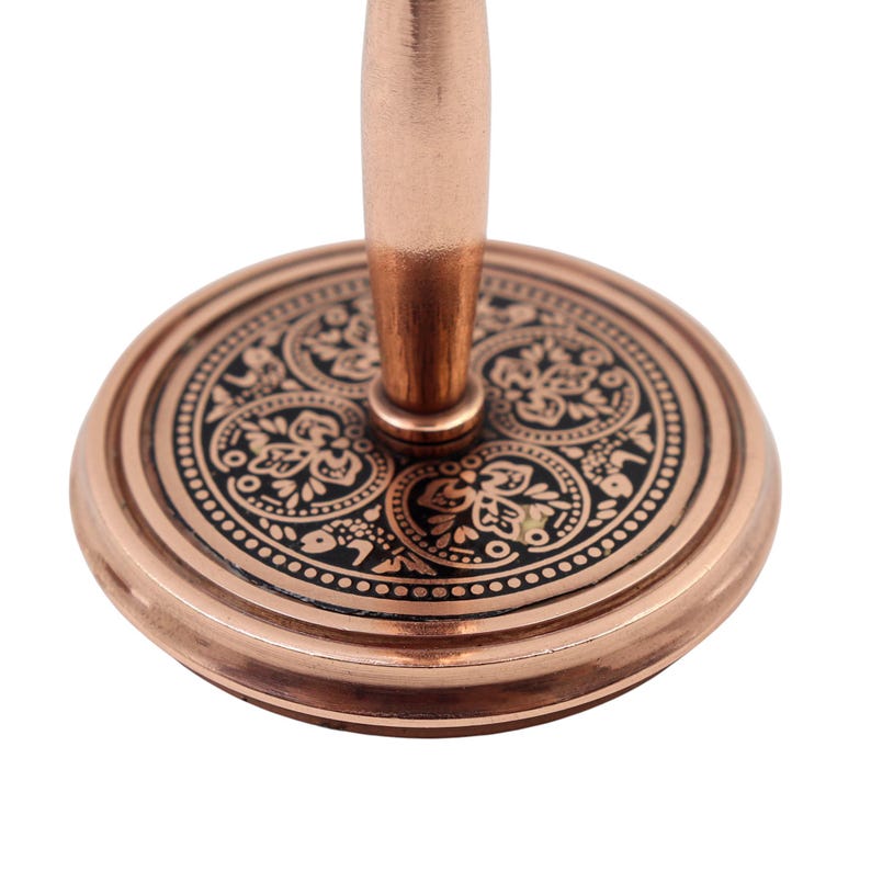 May include: A copper-toned metal stand featuring a circular base with a black inlay. The base is decorated with intricate floral and leaf patterns in copper. A smooth, cylindrical copper-toned post extends vertically from the center of the base.