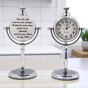 May include: Two silver-toned pocket watch clocks on stands. One clock face displays the time, the other has an inscription: "Time has only made our love stronger. Twenty five years down. forever to go. Charlotte with love from Oliver 14 Aug 1999."