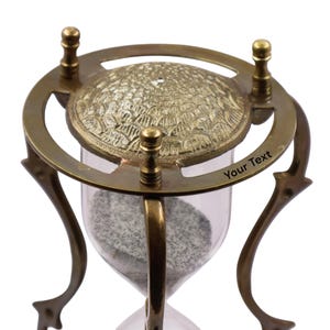Puede incluir: Un reloj de arena con marco de lat&oacute;n, patas curvas y una placa superior circular con un patr&oacute;n texturizado de escamas. El marco de lat&oacute;n incluye tres remates verticales y el texto grabado Your Text en el anillo circular superior.
