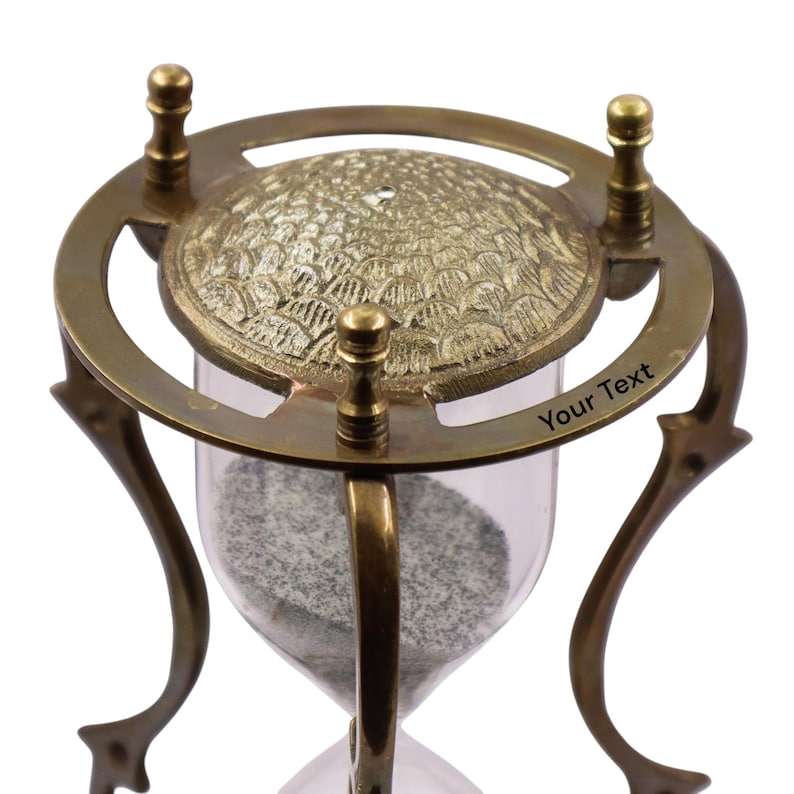 Puede incluir: Un reloj de arena con marco de lat&oacute;n, patas curvas y una placa superior circular con un patr&oacute;n texturizado de escamas. El marco de lat&oacute;n incluye tres remates verticales y el texto grabado Your Text en el anillo circular superior.