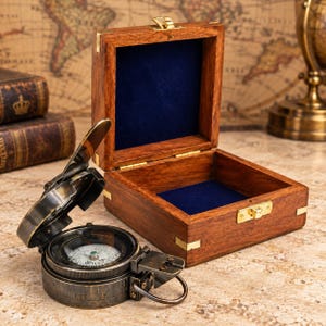 Peut inclure: Une boîte en bois brun foncé avec un couvercle à charnière et une doublure intérieure en velours bleu profond, dotée d'un loquet en laiton. À côté de la boîte se trouve une boussole ronde en laiton de style ancien avec un couvercle.