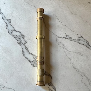 May include: A vintage brass telescope with a telescoping design. The brass has a warm, golden hue, and the telescope is composed of multiple cylindrical sections. The base and top have a slightly wider diameter than the central sections. The telescope is approximately 25 cm long.