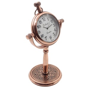 May include: A copper-toned metal desk clock with a circular face, white dial, and black Arabic numerals. The clock features the text Dollond London on the face, a winding crown at the top, and a decorative circular base with an intricate pattern.
