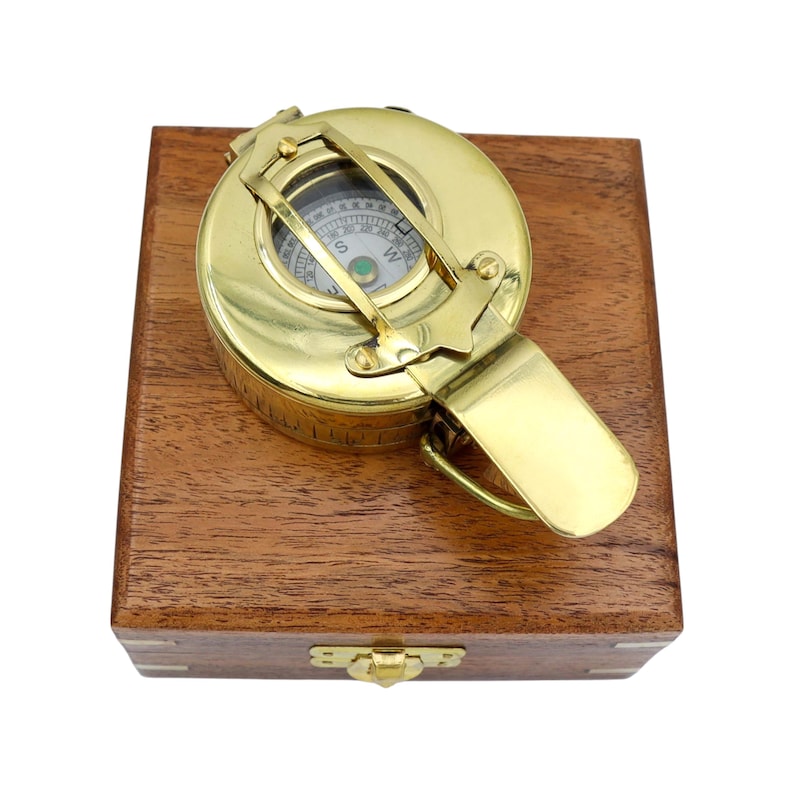 May include: A polished brass nautical compass with a hinged lid and protective metal bars over the glass face, resting on top of a square, dark brown wooden storage box featuring a brass latch closure on the front side of the container.