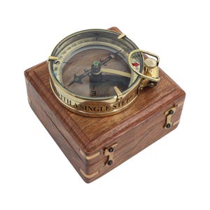 May include: A brass nautical compass with a glass top sits atop a square wooden box. The compass features a needle, directional markings, and the engraved text "WITH A SINGLE STEP" along its side. The box has brass corner accents and hinges.
