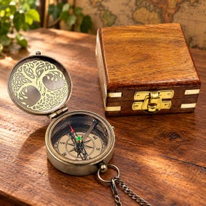 May include: A brass pocket compass with a tree design engraved on the interior lid, featuring a detailed directional dial with a red-tipped needle. The compass is accompanied by a square, polished wooden storage box with brass metal hardware.