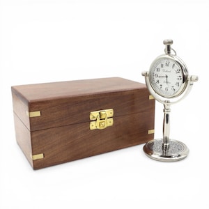 May include: A silver pocket watch on a stand beside a wooden box. The watch has a white face with black numerals and hands. The box is brown with brass accents and a gold clasp. The watch is approximately 10 cm tall.