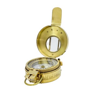 May include: A circular brass compass with a hinged lid featuring an integrated viewing window. The base displays a detailed directional dial with numerical markings and a small metal loop attached to the side for secure handling or attachment.