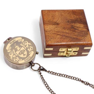 May include: A vintage brass compass featuring a family crest and the name "Boyle". The compass is on a chain, next to a wooden box with brass detailing. The box has a clasp.