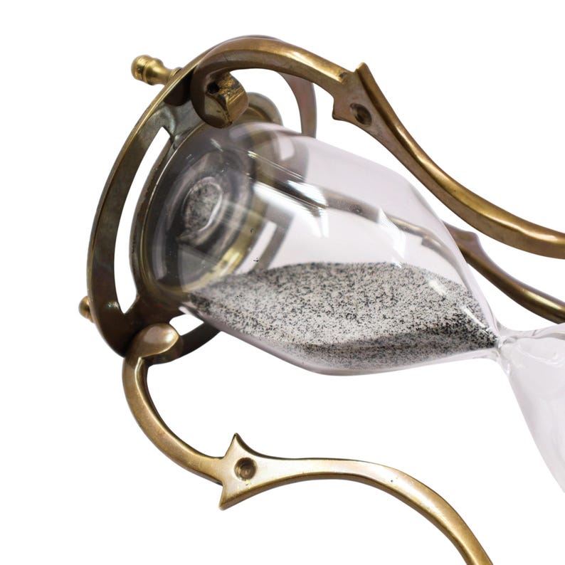 Puede incluir: Un reloj de arena con un marco de lat&oacute;n pulido con soportes curvos y ornamentados y un bulbo de vidrio transparente que contiene arena fina de color gris oscuro. La estructura met&aacute;lica conecta las secciones superior e inferior.