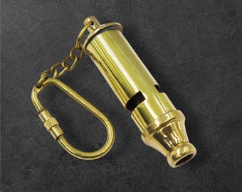 May include: A polished brass whistle featuring a cylindrical body with a side sound aperture and a flared mouthpiece. The whistle is attached to a short metal chain connected to a screw-lock carabiner, all finished in a matching brass tone.