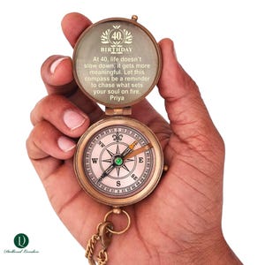 Peut inclure: Une boussole vintage en laiton avec un couvercle à charnière, ouverte pour révéler une rose des vents et du texte gravé. Le couvercle indique "40 Anniversaire" et un message personnalisé. Une chaîne et le logo Dollond London sont visibles.