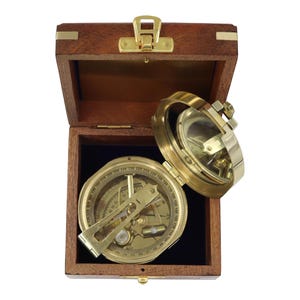 May include: A brass nautical compass with a hinged lid and internal dial featuring numerical markings, resting inside a square dark brown wooden box with a gold-toned metal latch and hinges. The compass has a metallic finish and visible gears.