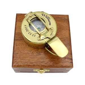 May include: A polished brass pocket compass with a hinged lid resting on a square wooden box. The compass lid is engraved with the name William and the date 12.12.2025. The compass features a visible directional dial and a metal latch mechanism.