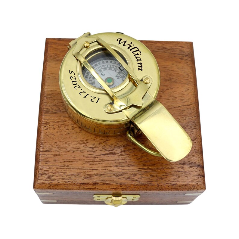 May include: A polished brass pocket compass with a hinged lid resting on a square wooden box. The compass lid is engraved with the name William and the date 12.12.2025. The compass features a visible directional dial and a metal latch mechanism.