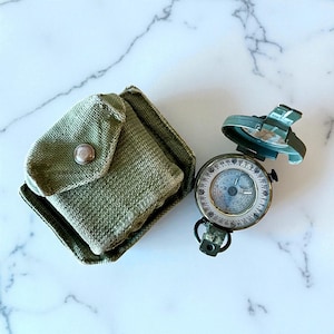 Puede incluir: Una brújula verde oliva con tapa abatible y funda de lona a juego. La esfera de la brújula tiene un fondo blanco con marcas negras y rojas. La funda tiene un cierre de botón a presión.
