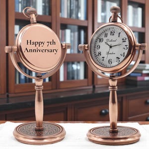 May include: Two copper-coloured, vintage-style clocks. One clock face reads "Happy 7th Anniversary." The other clock face reads "Dollond London." Both clocks have a round face, a stand, and a decorative base. The clocks are displayed on a white surface.