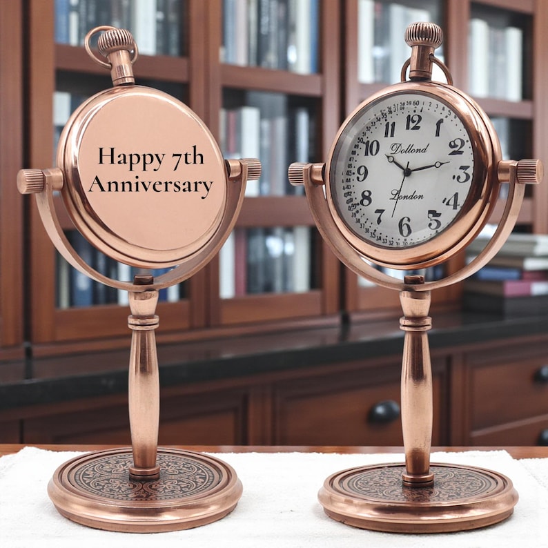 May include: Two copper-toned desk clocks on circular bases. The left clock features the text "Happy 7th Anniversary" on its face. The right clock has a white dial with black numerals, the text "Dollond London," and a winding crown at the top.