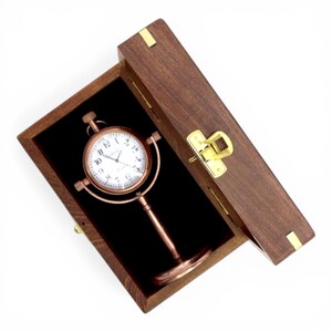 May include: A copper-toned gimbal clock with a white face and black numerals, mounted on a pedestal stand. The clock is housed inside a rectangular wooden box with a brass-colored latch and hinges, featuring a black interior lining.