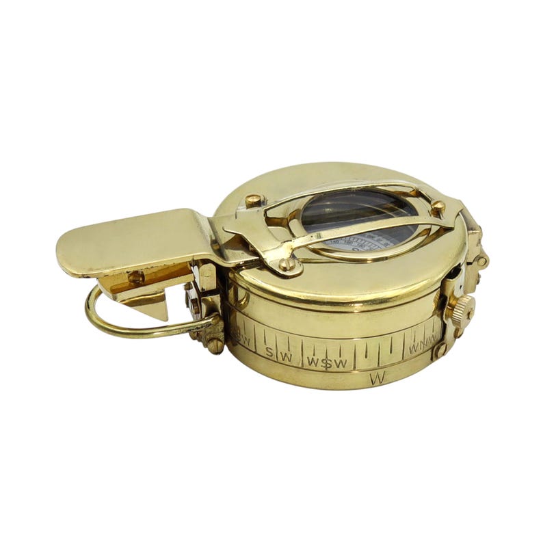 May include: A polished brass pocket compass featuring a circular casing with a hinged lid, a protective metal bridge over the glass face, and a side-mounted adjustment knob. The outer rim is engraved with directional markings including W, WSW, and SW.
