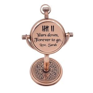 May include: A copper-toned metal desk clock with a circular face featuring the text '7 Years down, Forever to go. Love, Sarah' and a tally mark graphic. The clock is mounted on a pedestal base with an intricate floral pattern and a top ring.