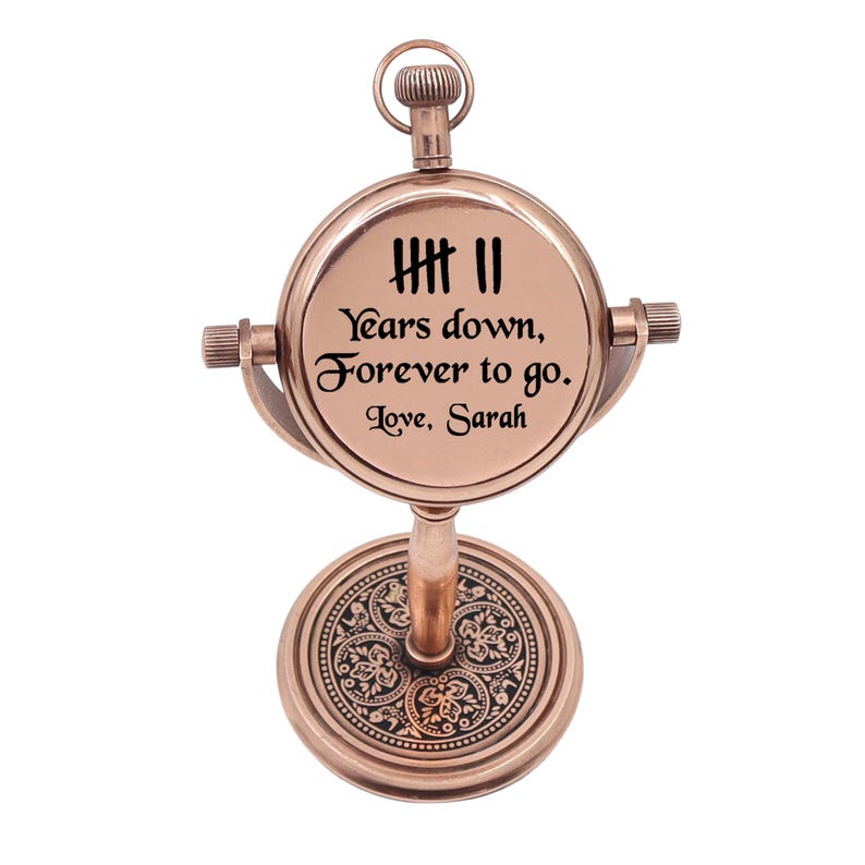 May include: A copper-toned metal desk clock with a circular face featuring the text '7 Years down, Forever to go. Love, Sarah' and a tally mark graphic. The clock is mounted on a pedestal base with an intricate floral pattern and a top ring.