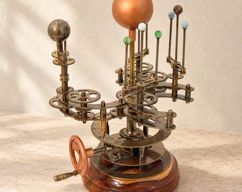 May include: A mechanical orrery model featuring a large central copper-colored sphere representing the sun, surrounded by smaller planetary spheres in various colors and textures, all mounted on a dark wooden base with brass gears and a hand crank.
