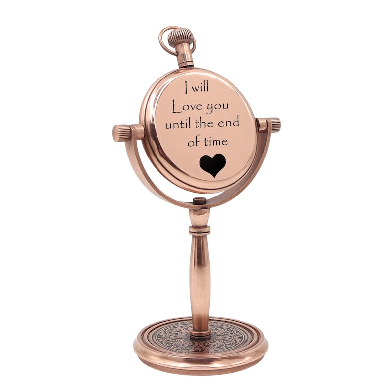 May include: A copper-toned metal desk clock on a pedestal stand. The circular clock face features the engraved text 'I will Love you until the end of time' above a black heart icon. The base has an intricate circular pattern engraved on it.