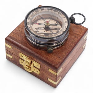 May include: A vintage compass with a black metal casing and a clear glass top, resting on a wooden box. The compass face features directional markings and a needle. The box is made of dark wood with brass corner accents and a clasp.
