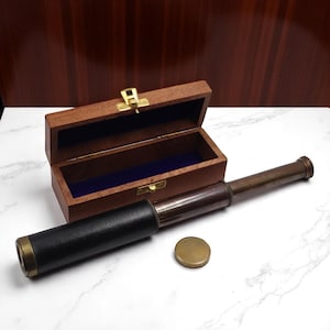 Peut inclure: Une longue-vue portative en laiton gainée de cuir marron foncé avec un capuchon d'objectif en laiton assorti. La longue-vue est placée devant une boîte de rangement rectangulaire en bois ouverte avec un fermoir en métal doré et un intérieur en velours bleu.