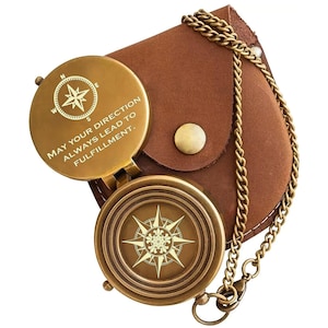 May include: A brass pocket compass with a hinged lid featuring the engraved text MAY YOUR DIRECTION ALWAYS LEAD TO FULFILLMENT and a compass rose design. The compass is attached to a gold-toned metal chain and rests on a brown leather pouch.