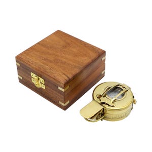 May include: A polished brass compass with a hinged lid and a visible directional dial, positioned next to a square wooden storage box featuring brass corner accents and a central brass latch mechanism on the front side of the container.