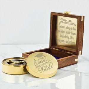 Peut inclure: Une boîte en bois avec une plaque en laiton à l'intérieur et une boussole et un couvercle dorés. Le couvercle indique "The Tassel Was Worth The Hassle" avec un graphisme de chapeau de graduation. La plaque de la boîte indique "Happy Graduation".