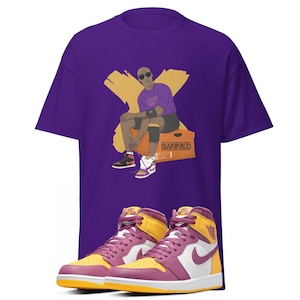 Puede incluir: Camiseta morada con un gráfico de un jugador de baloncesto que lleva una camiseta morada y amarilla y está sentado en una caja que dice "Banned". La camiseta se combina con un par de zapatillas moradas, amarillas y blancas.