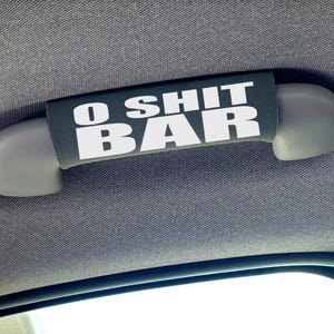 Puede incluir: Una funda rectangular negra para la manija de agarre del coche con el texto blanco O SHIT BAR impreso en una fuente sans-serif en negrita. La funda está envuelta alrededor de una manija de techo gris del vehículo.