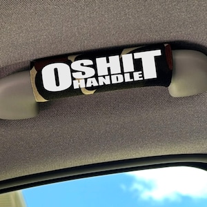 May include: A camouflage patterned fabric cover for a vehicle grab handle, featuring the white text O SHIT HANDLE printed across the centre. The cover is wrapped around a light grey plastic handle mounted to the interior ceiling of a car.